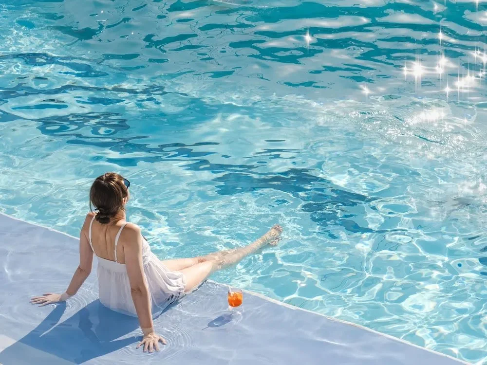 A woman with short brown hair in a white dress sitting on the edge of a swimming pool with her legs in the water, a drink placed nearby, on a sunny day.