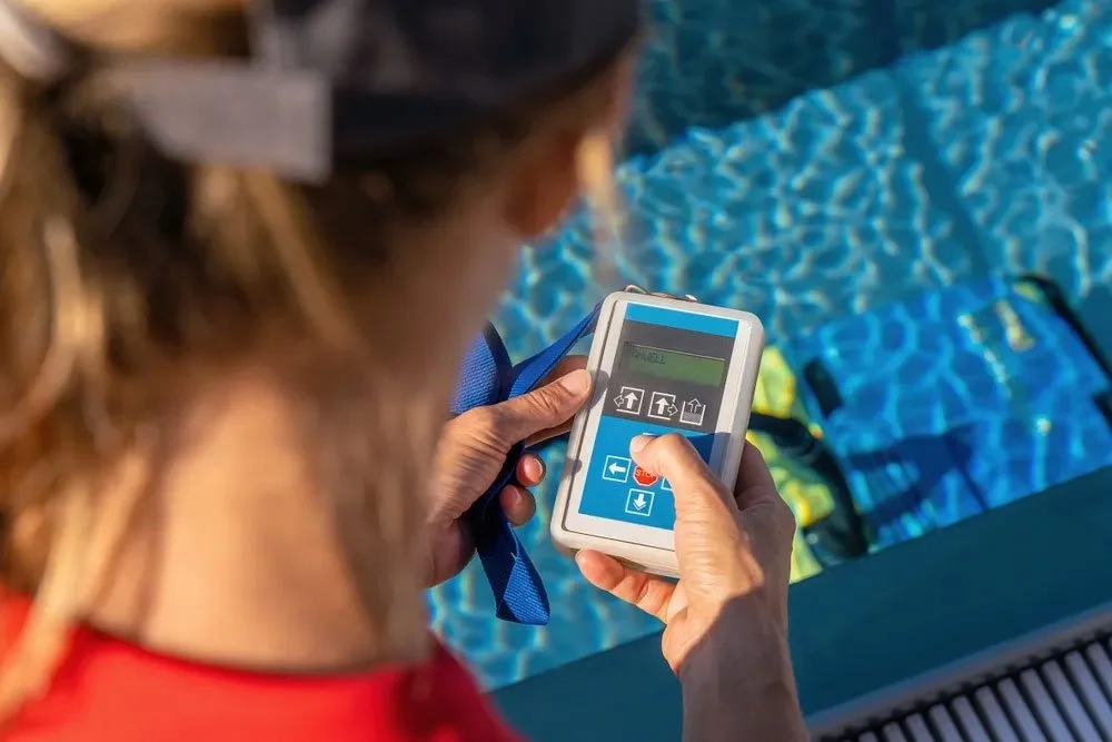 Person holding a remote control device with buttons, near a swimming pool with blue water and colorful pool floaties.