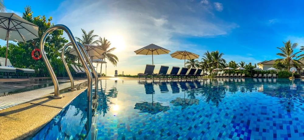 Empty swimming pool with underwater mosaic tiles, poolside chairs with umbrellas, palm trees, and a blue sky during sunset.