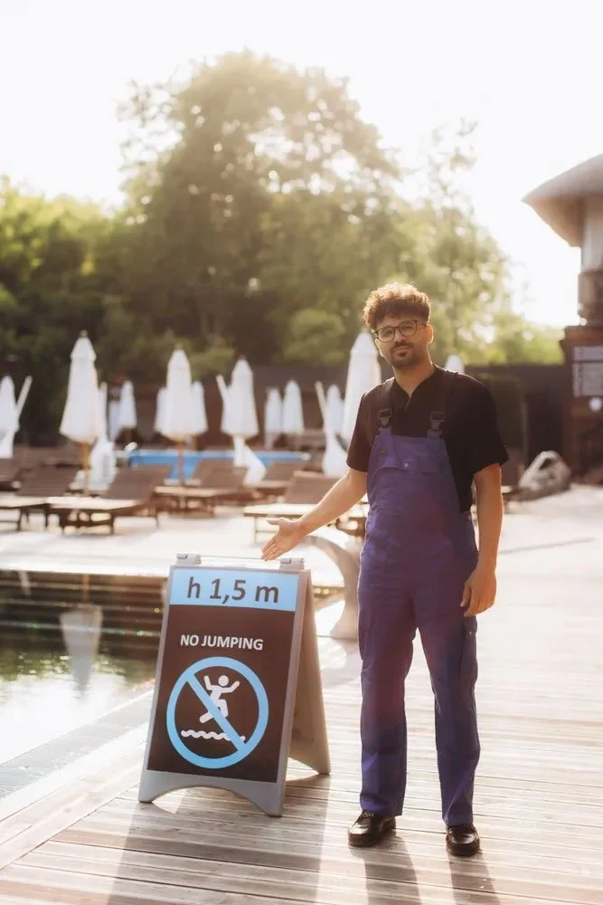 A man with curly hair and glasses wearing a black shirt and blue overalls standing on a wooden poolside deck, pointing to a sign that indicates a maximum height limit of 1.5 meters and no jumping, near a pool with a calm water surface, surrounded by lounge chairs and white umbrellas, with trees and a building in the background.