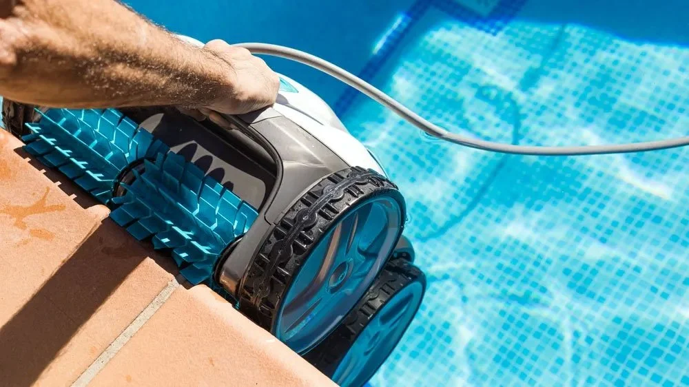 A robotic pool cleaner scrubbing the edge of a swimming pool with blue tiles.