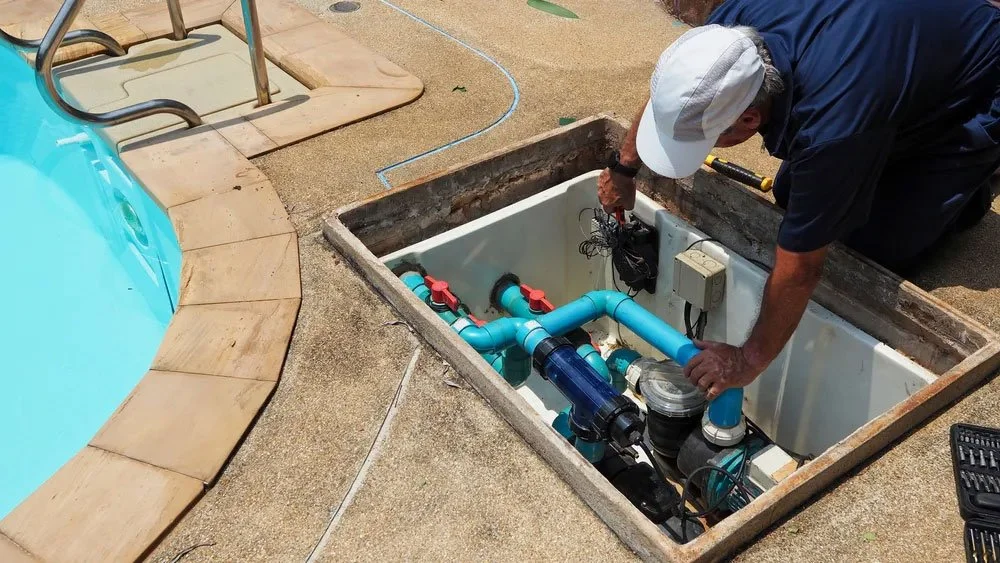 A worker installing or repairing pool equipment inside an underground access panel next to a swimming pool.
