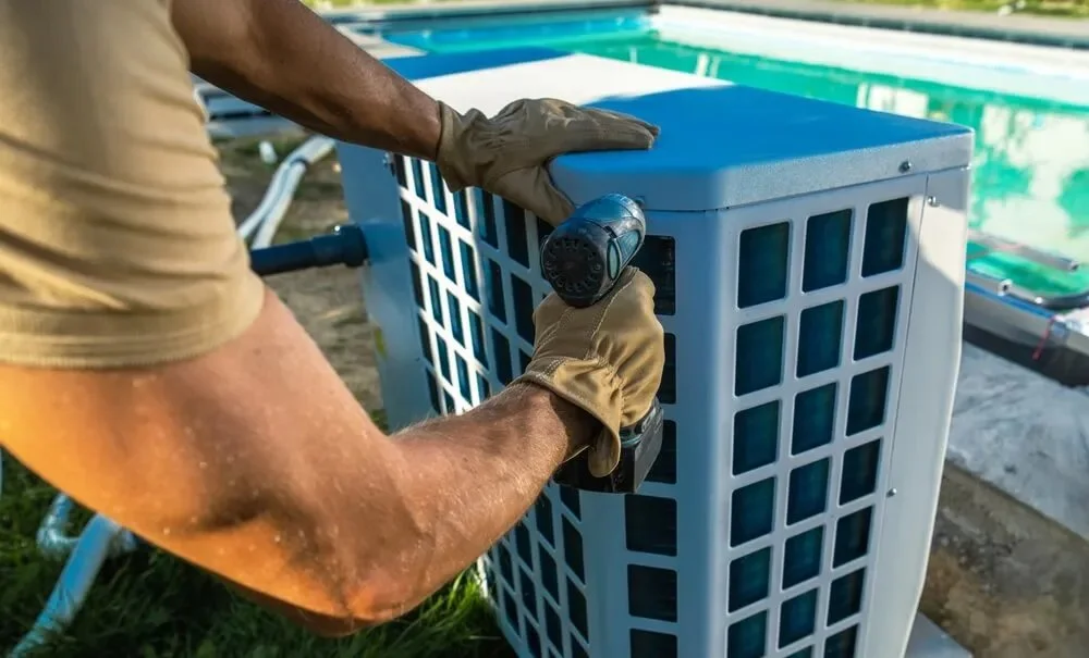 A person in gloves using a power drill to install or repair an outdoor air conditioning unit next to a swimming pool.
