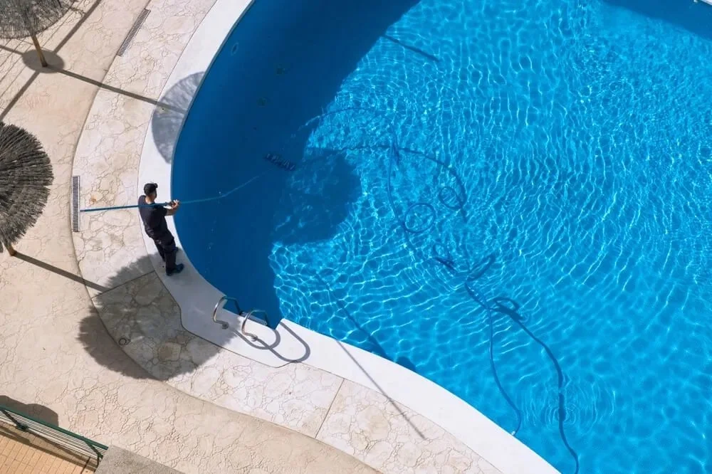 Worker cleaning a large blue swimming pool with a telescopic pole in a backyard.