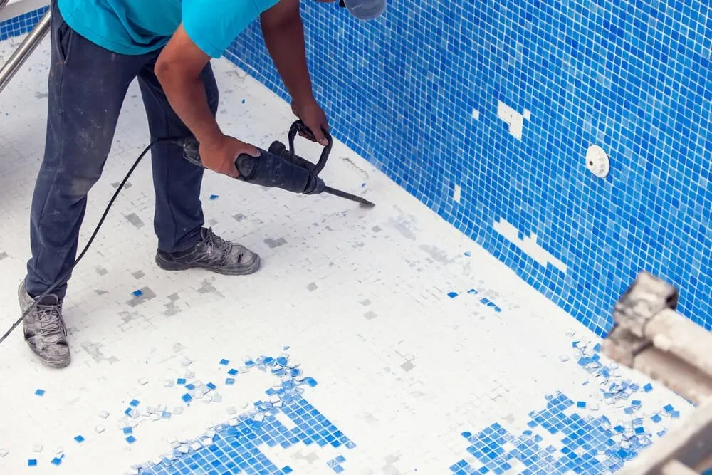 A person in a teal shirt and jeans is using a power tool to remove blue mosaic tiles from a pool wall, with broken tiles scattered on the pool floor.