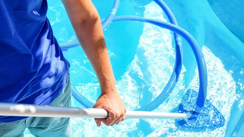 A person in a blue shirt cleaning a swimming pool with a pool vacuum cleaner.