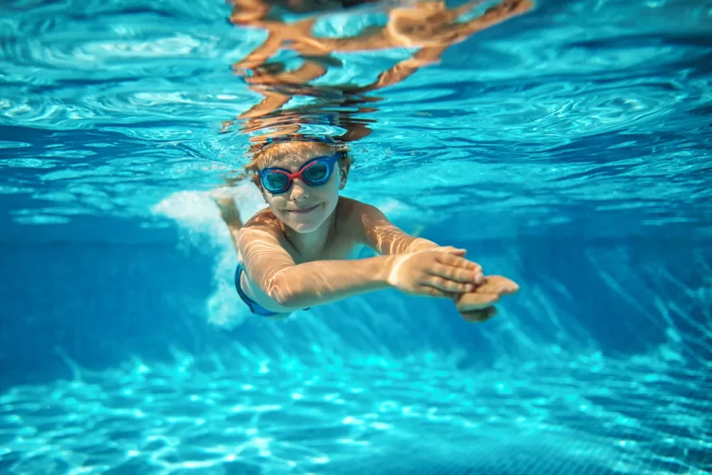 Child swimming underwater in a pool wearing blue goggles and smiling.
