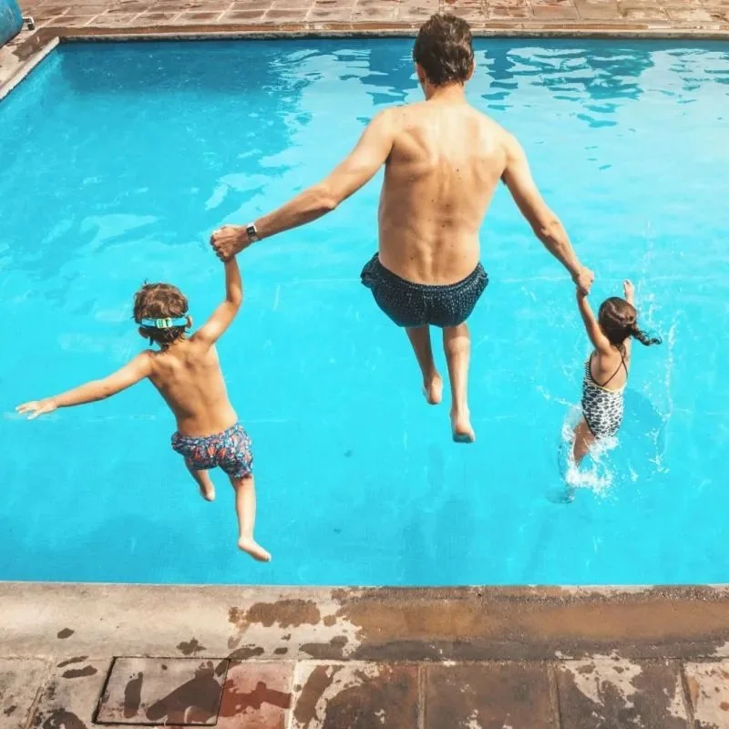 A man and two children jumping into a swimming pool, holding hands, with the man in the center and the children on either side.