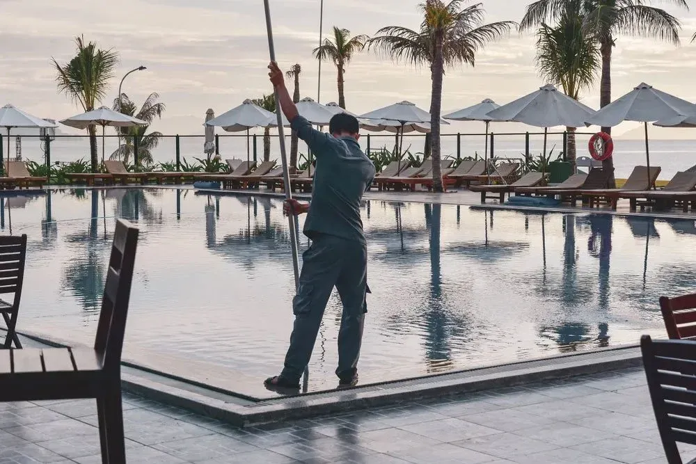 A man cleaning a swimming pool at a tropical resort with palm trees and lounge chairs.